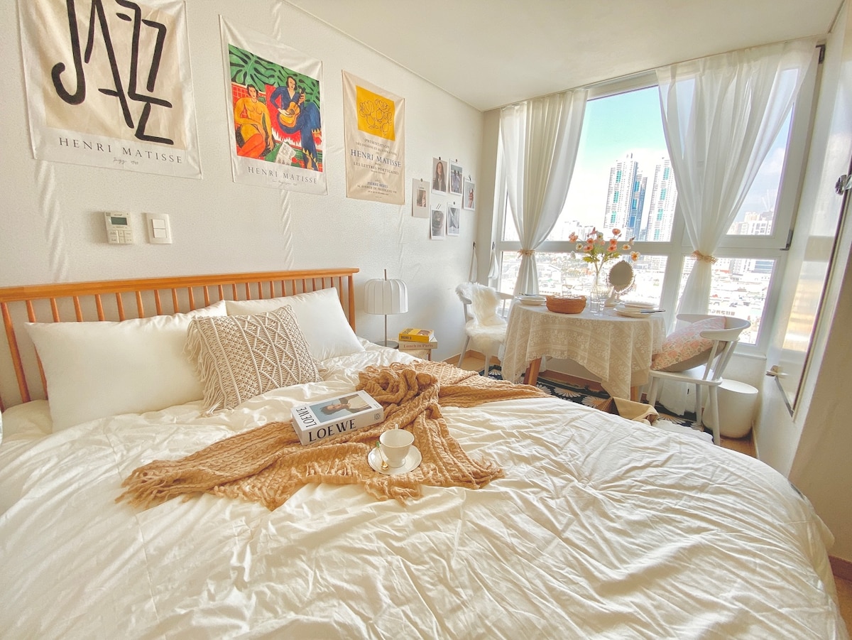A bright and airy bedroom features a spacious bed adorned with a cozy throw and decorative pillows. The window offers a city view, while soft natural light illuminates the space. A small table is arranged nearby, accompanied by a chair and personal touches like wall art.