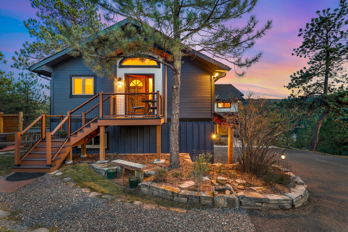A welcoming entryway is framed by natural landscaping, featuring a curved stone path leading to a wooden deck. Large windows illuminate the exterior, reflecting the colors of sunset. A comfortable chair sits on the porch, inviting relaxation amid the tranquil surroundings.