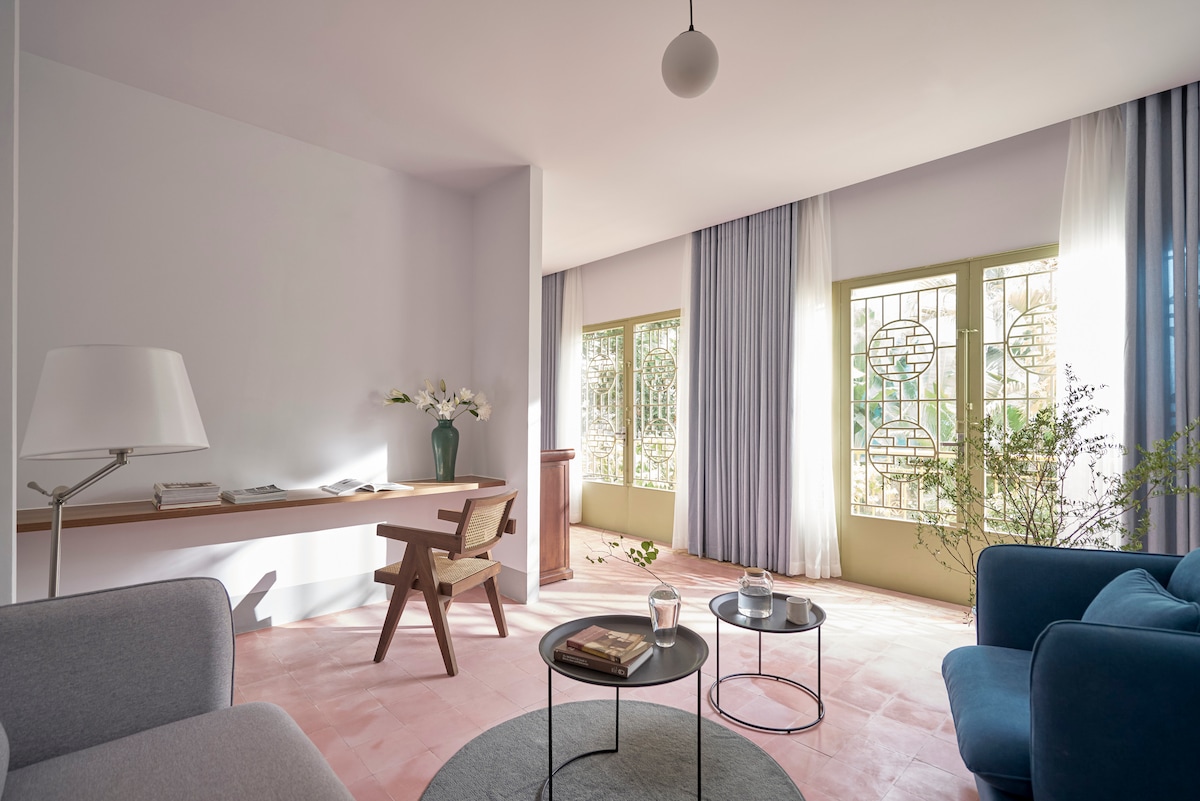 A light-filled living space features large windows with decorative grills, allowing natural light to illuminate the room. A simple wooden table is paired with an armless chair, while a soft blue sofa and a round grey rug add comfort to the area.