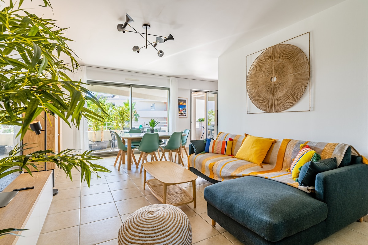 A spacious living area features a comfortable sectional sofa adorned with colorful cushions. A round coffee table sits in front, surrounded by modern dining chairs. Large glass doors lead to a terrace with greenery, while natural light brightens the room.