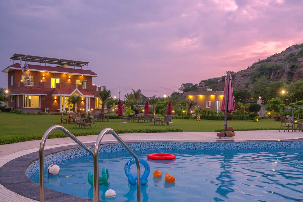 A private swimming pool is seen in the foreground, featuring colorful floating toys. The well-maintained garden surrounds the pool, dotted with seating areas and umbrellas, while the two-story red brick house stands in the background against a softly lit sky.