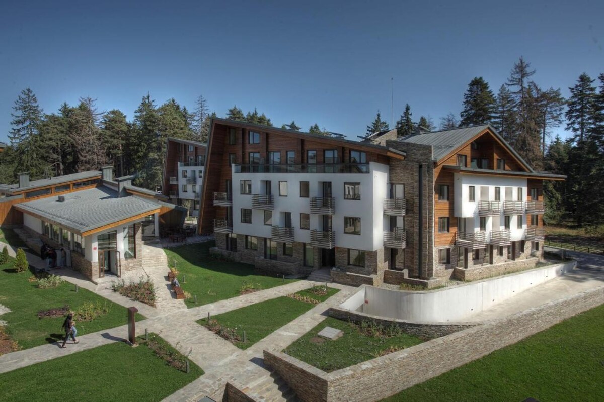 An expansive view of a modern hotel structure surrounded by lush greenery. The building showcases a blend of stone and wood elements, with multiple balconies and large windows. Well-maintained pathways meander through manicured lawns, enhancing the tranquil outdoor environment.