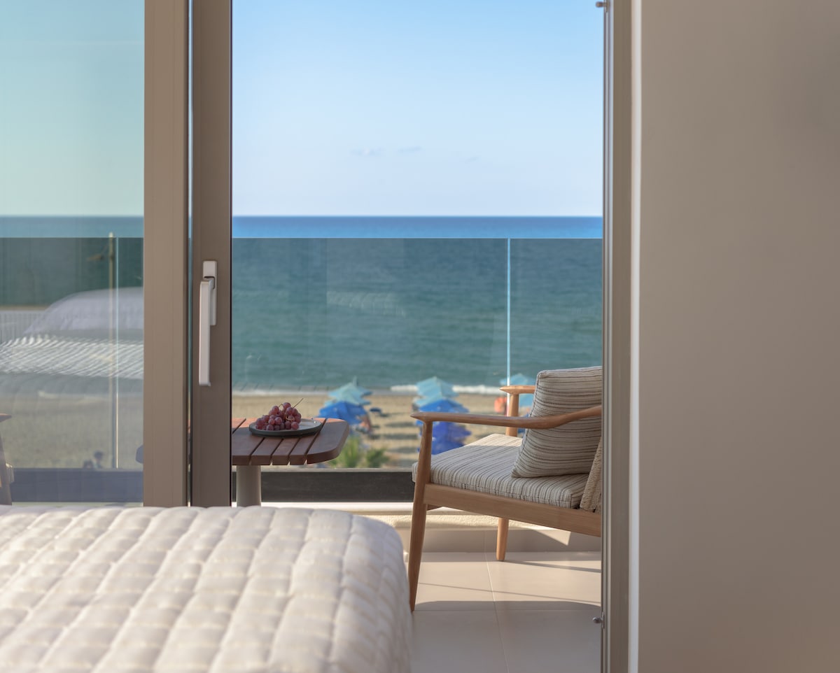 The image showcases a serene seafront view through glass doors, revealing the tranquil Mediterranean Sea. A cozy chair sits adjacent to a small table, complementing the inviting atmosphere. The beach with sun umbrellas can be seen in the distance, enhancing the coastal ambiance.