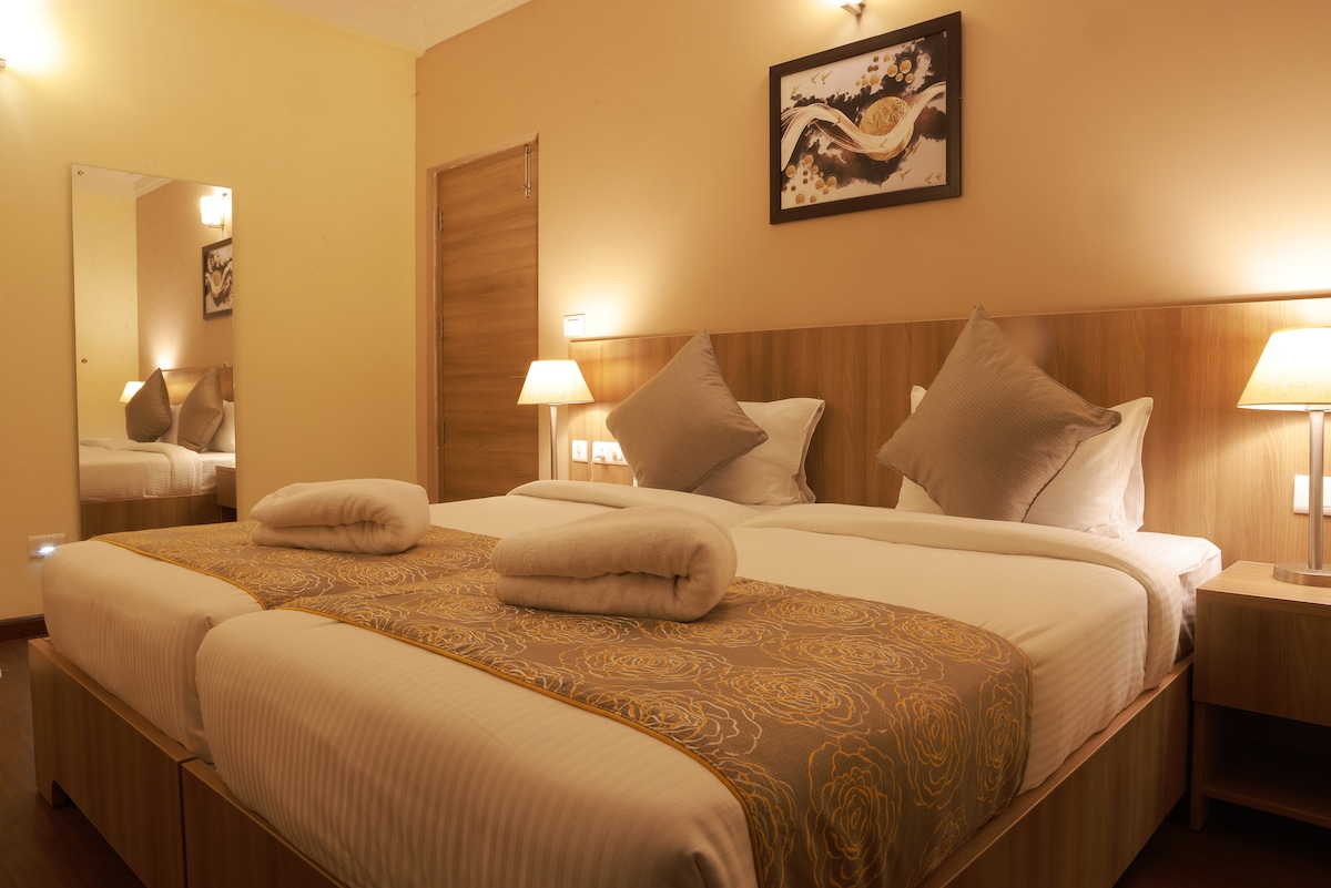 A well-appointed sleeping area features two spacious beds with tasteful linens and decorative cushions. Soft lighting from bedside lamps creates a relaxing ambiance, while a mirror on the wall enhances the sense of space. A piece of wall art adds a touch of character.