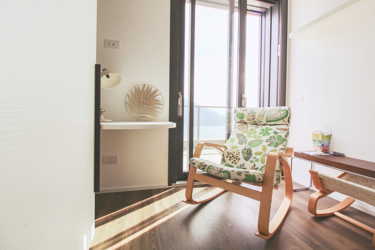 A rocking chair with a floral-patterned cushion is positioned beside a small wooden table. Natural light fills the space through large glass doors that open to a view of the surrounding landscape, creating a peaceful and inviting corner.