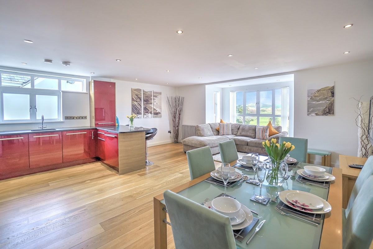 An open-plan living area features a modern kitchen with red cabinets and a dining table set for five. A spacious sectional sofa is positioned near large windows, welcoming natural light and offering views of the surrounding landscape. Subtle decor enhances the atmosphere.