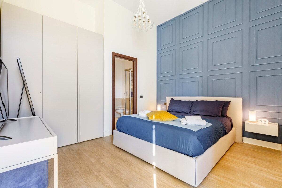 The bedroom features a spacious bed with dark bedding, complemented by vibrant accent pillows. A stylish blue accent wall adds depth to the room. A sleek closet stands nearby, and a modern chandelier hangs from the ceiling, providing warm illumination.