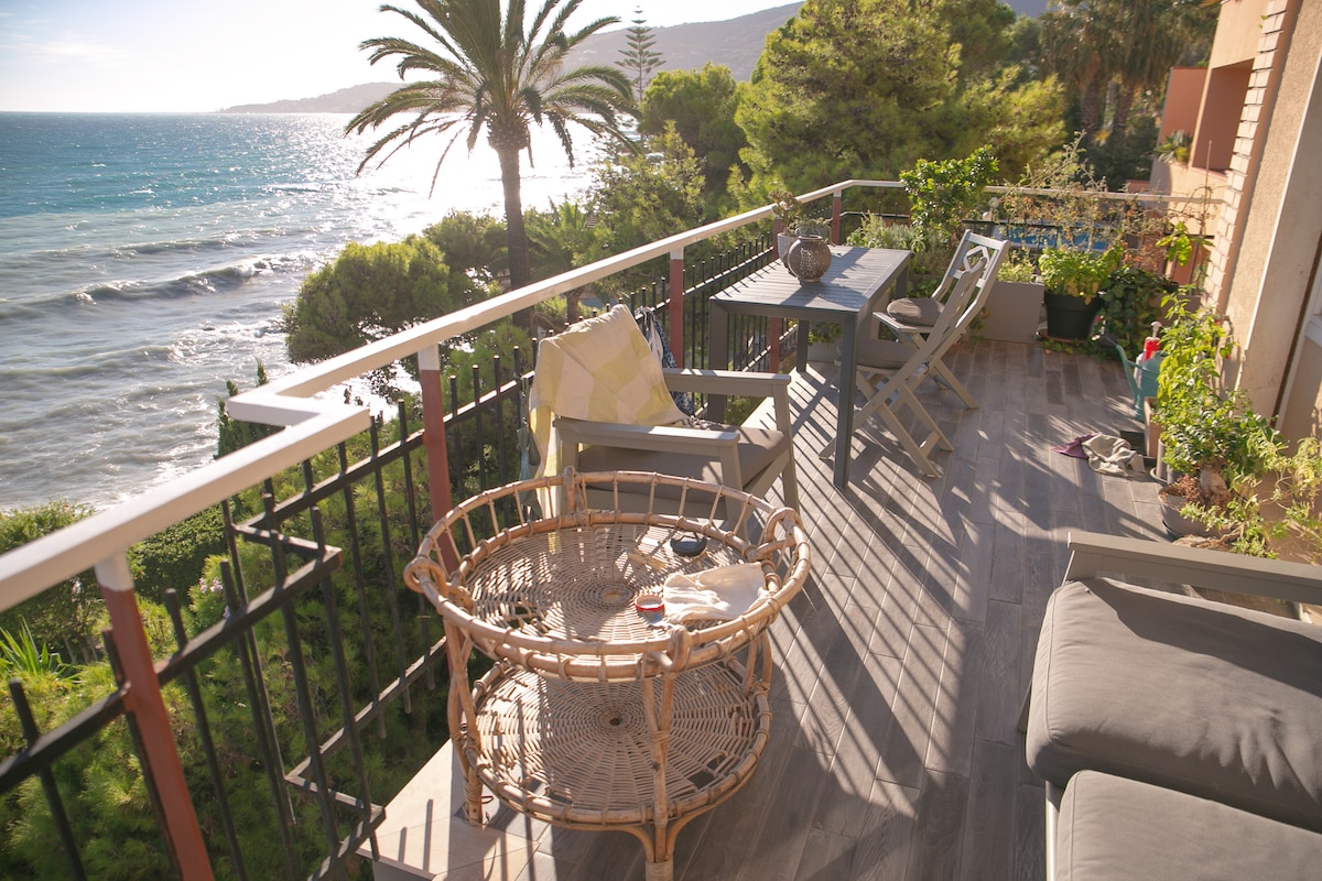 A spacious balcony overlooks the sea, featuring a comfortable seating area with a rattan chair and a table with two chairs. Lush greenery frames the view, while palm trees sway in the gentle breeze. The sound of waves can be imagined in this tranquil setting.