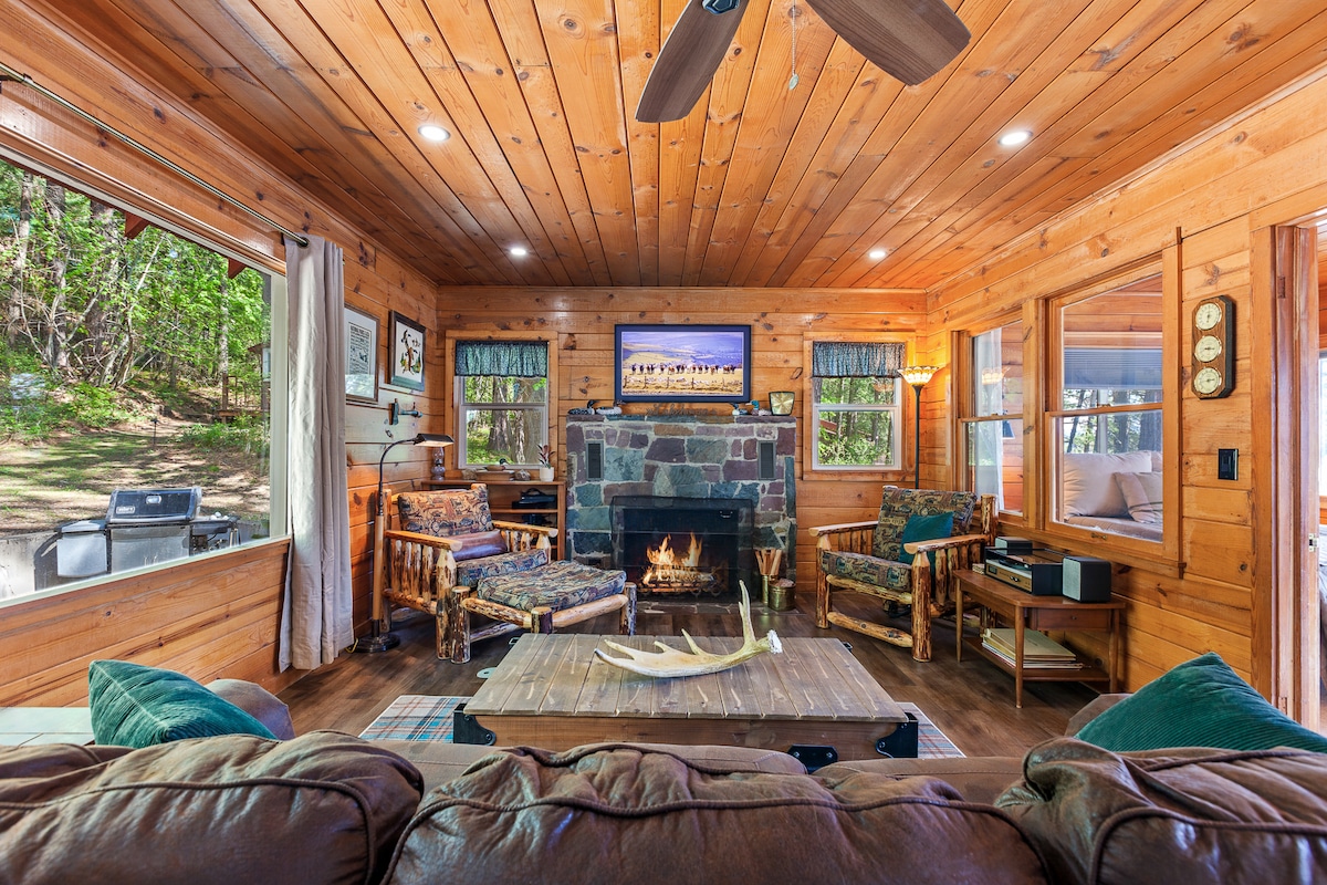 A warm living area features cedar-lined walls and ceiling, filled with light from large windows. Comfortable seating arrangements include rustic chairs and a plush couch facing a stone fireplace. A wooden coffee table sits at the center, with a television mounted above the mantel.