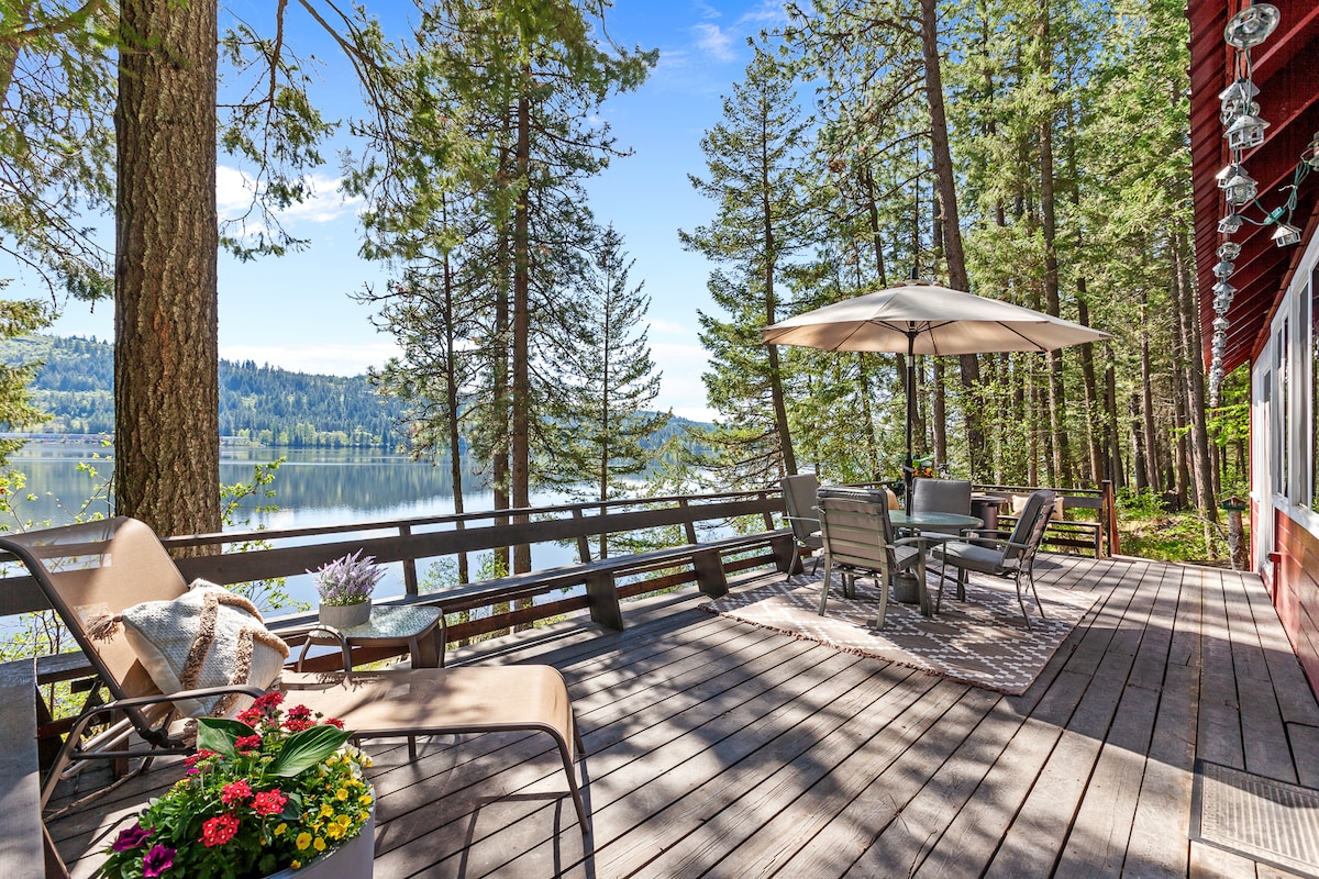 A spacious deck offers a tranquil view of the lake, surrounded by tall trees. Outdoor seating is arranged around a large table, shaded by a patio umbrella. A colorful planter adds a touch of greenery, enhancing the inviting atmosphere of the outdoor space.