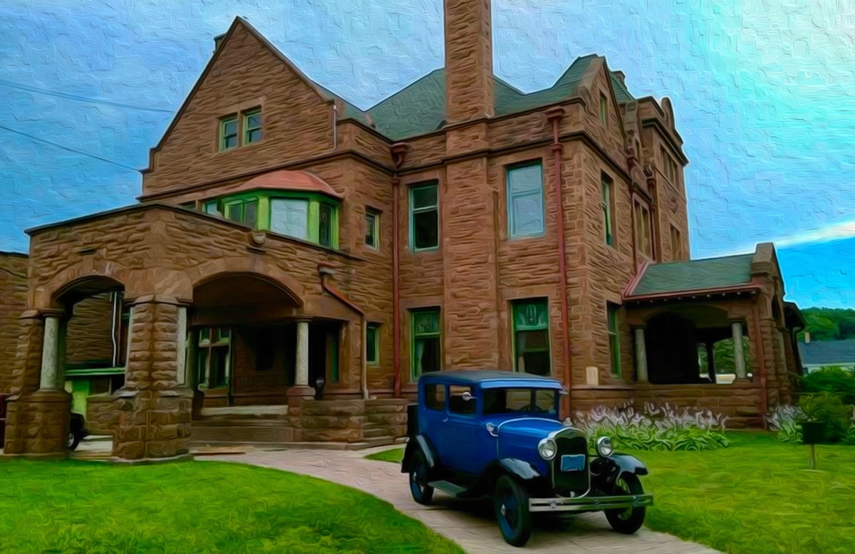 A vintage blue car is parked in front of a historic mansion, featuring a unique stone façade. The mansion's architecture includes prominent gables and multiple chimneys. Lush green grass surrounds the path leading to the entrance, emphasizing the building's grandeur against a cloudy sky.