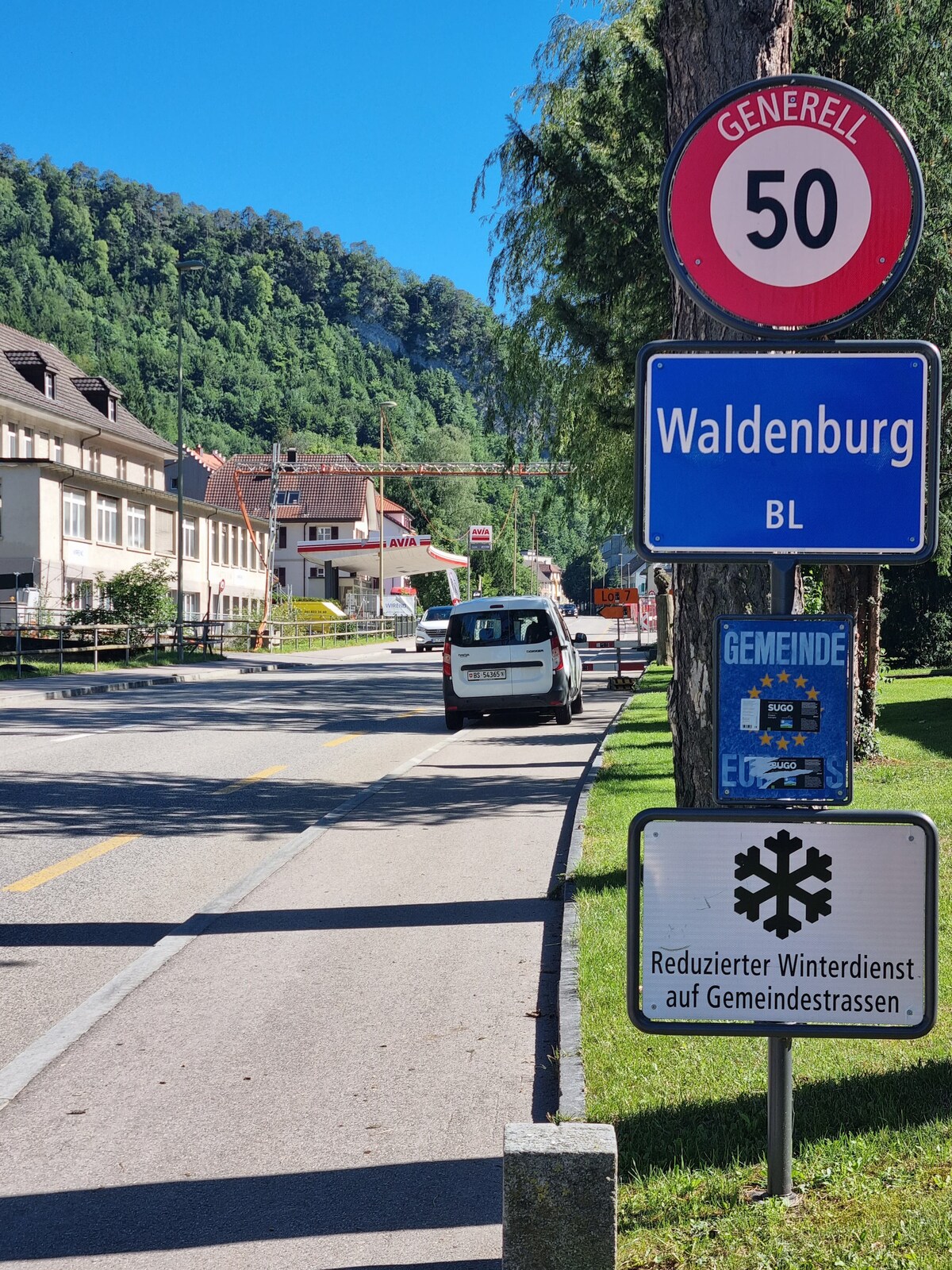 A roadside scene is captured, featuring a speed limit sign indicating 50 km/h alongside a blue sign marking the entry to Waldenburg. A white vehicle is parked nearby, with green trees lining the backdrop, creating a natural frame around the area.
