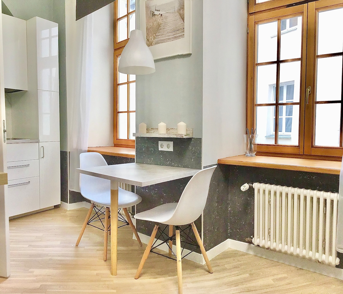 A dining area features a sleek table accompanied by two modern chairs, illuminated by a pendant lamp. Natural light enters through large windows with light curtains, highlighting the minimalist design. Wall-mounted candles add subtle warmth to the space.