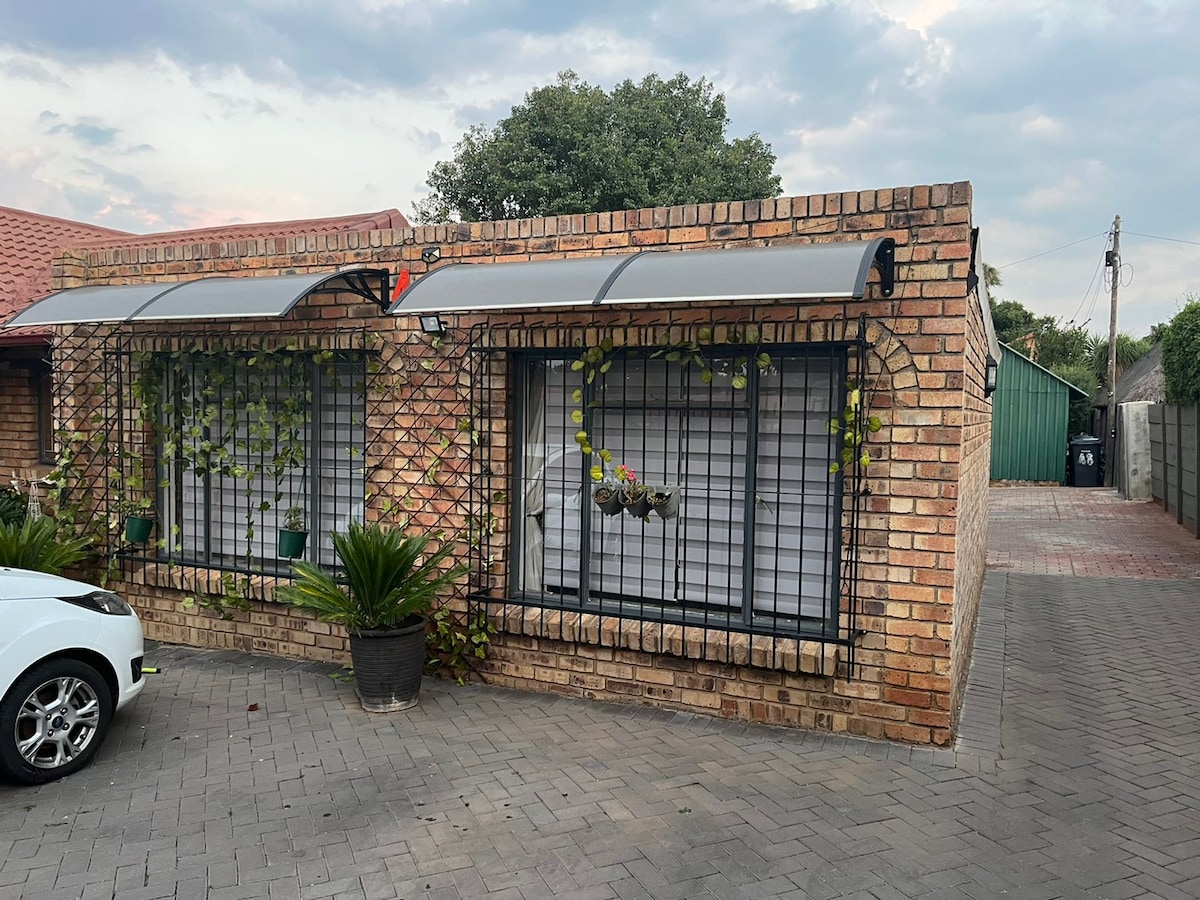 The exterior of a brick apartment is shown, featuring large windows adorned with decorative plant hangers and protective grilles. An awning extends over the windows. A vehicle is parked in front, and a paved driveway leads to additional greenery and a fenced area in the background.