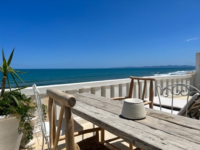 A wooden dining table is positioned on a sunlit terrace, overlooking the expansive sea. Nearby, a potted plant adds greenery to the setting. The tranquil blue waters and clear sky create a serene backdrop for outdoor meals.