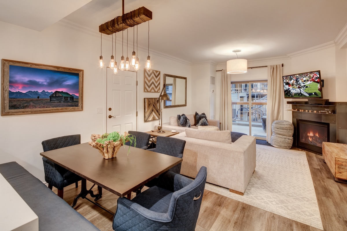 The open living area includes a dining table with dark chairs, a comfortable sofa, and a large flat-screen TV. Natural light fills the space through the windows, which offer views of the outdoors. A modern fireplace adds warmth to the ambiance.