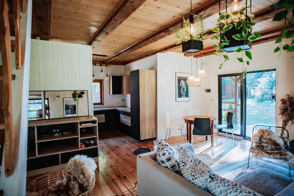 An open living space features wooden beams and a mix of natural light and greenery. A dining table with chairs is positioned near glass doors that lead to the outdoor area. A cozy sofa with decorative pillows is arranged around a textured rug, enhancing the inviting ambiance.