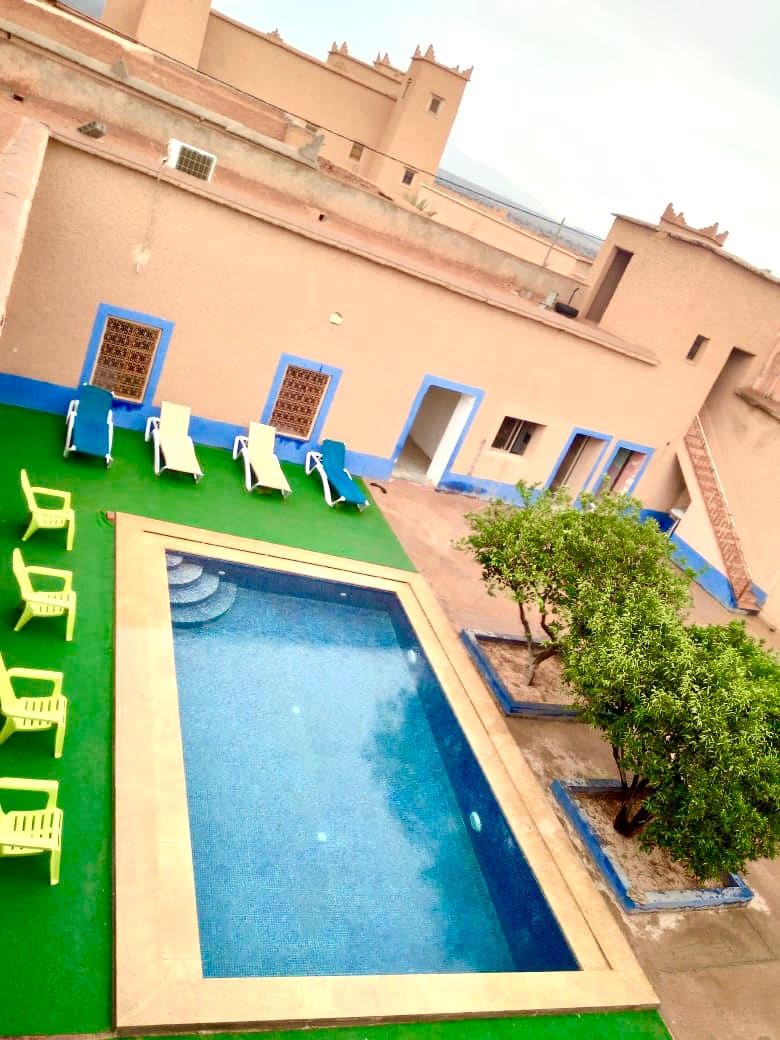 A serene outdoor area features a blue-tiled swimming pool surrounded by a green lawn. Sun loungers in vibrant yellow are arranged near the pool. The backdrop consists of a beige structure with blue accents, highlighting intricate architectural details.