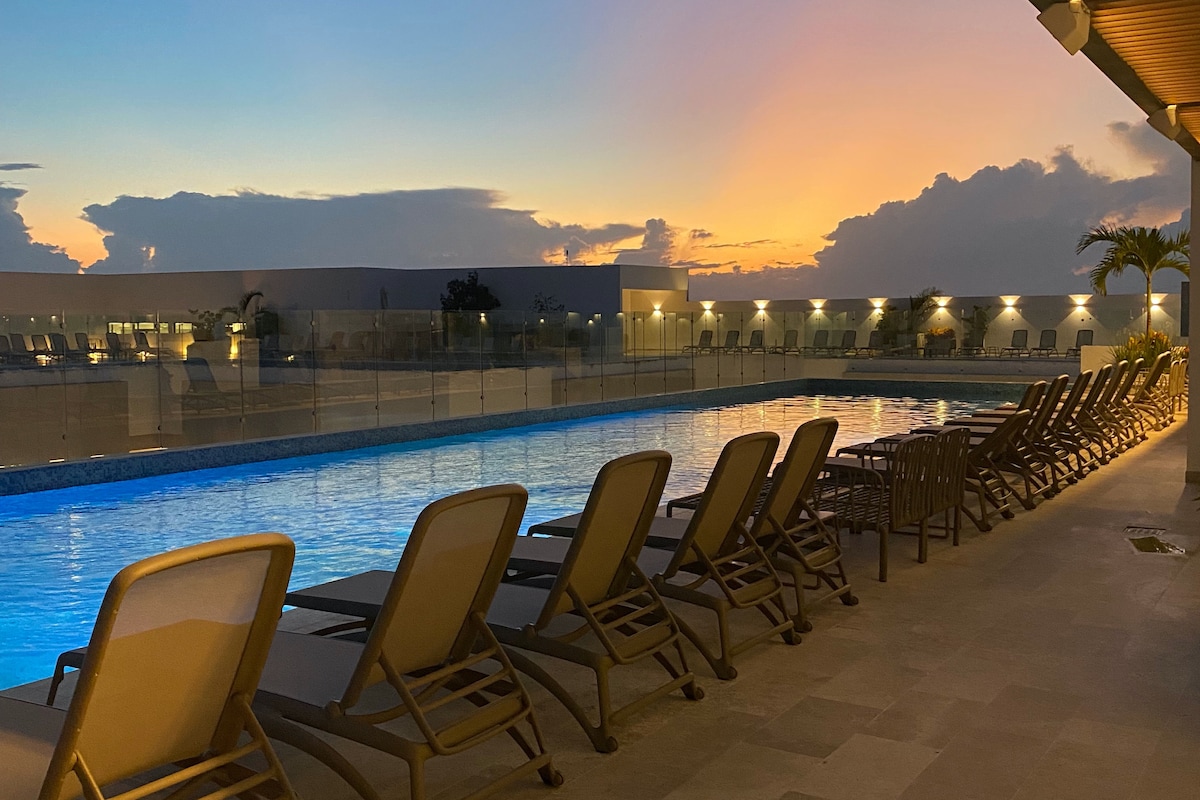 A serene rooftop pool area is showcased, featuring multiple lounge chairs arranged alongside the illuminated water. The setting is complemented by a soft sunset sky, reflecting hues of orange and blue, with palm trees gently framing the scene.
