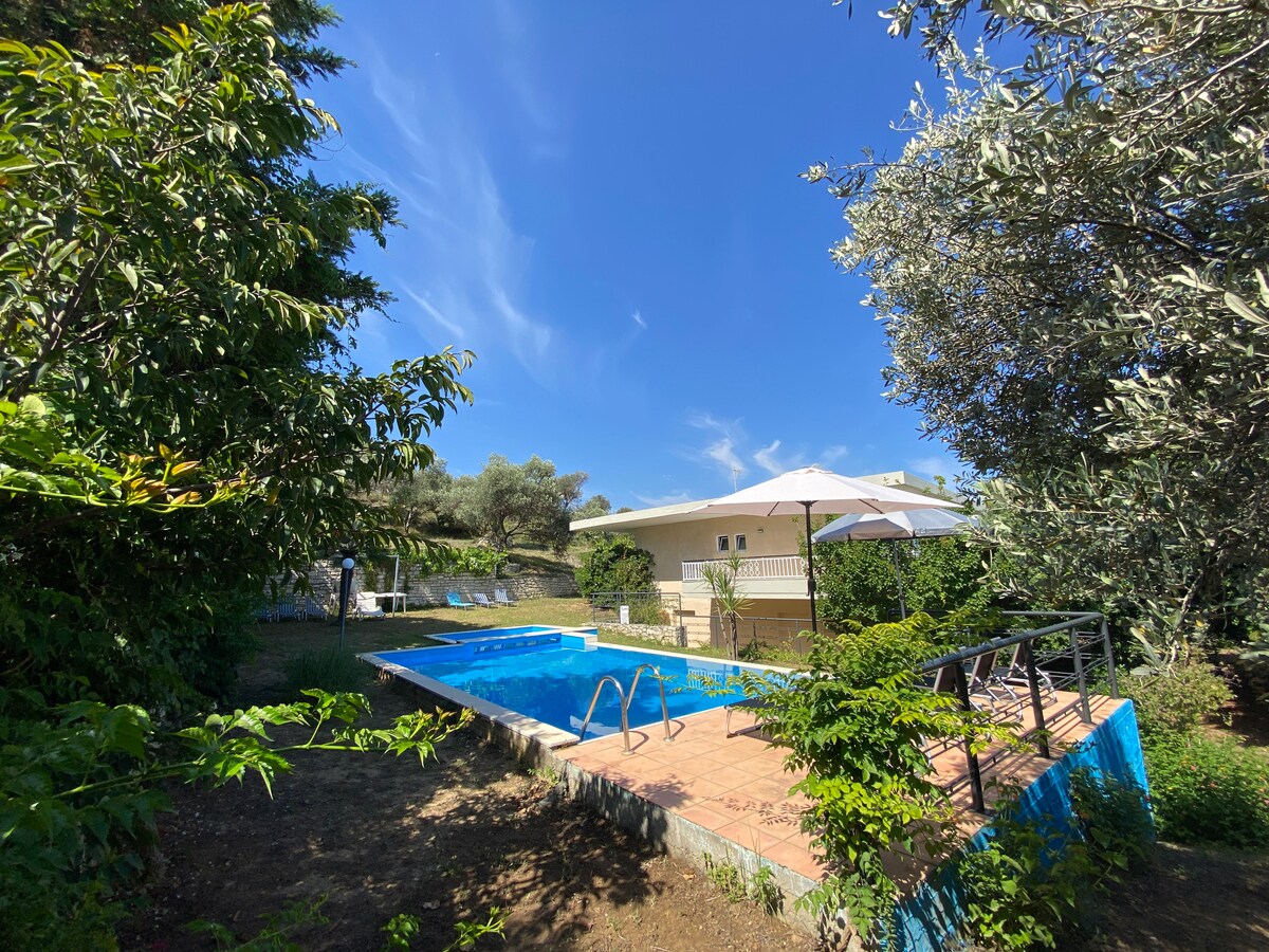 A swimming pool is visible, surrounded by lush greenery and trees. An umbrella offers shade on the pool deck, while a light-colored building peeks through the foliage in the background. The clear blue sky complements the tranquil setting.