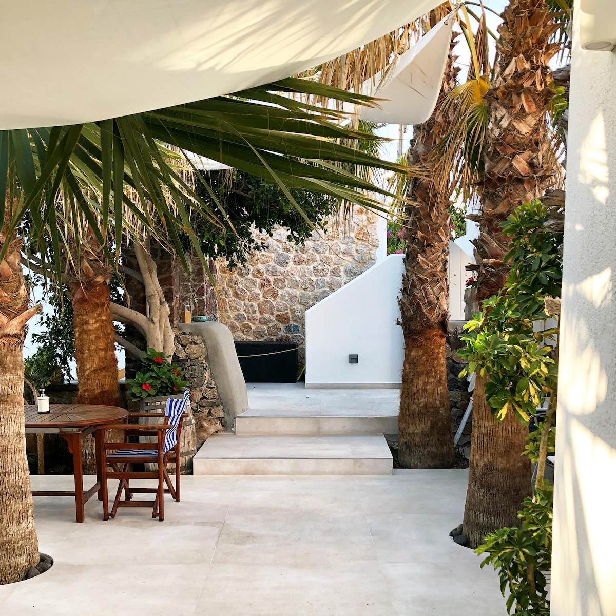 A serene outdoor area is framed by palm trees and greenery, providing a shaded space with a wooden table and chairs. Stone steps lead up to a natural stone wall, creating a relaxing atmosphere surrounded by lush plants.