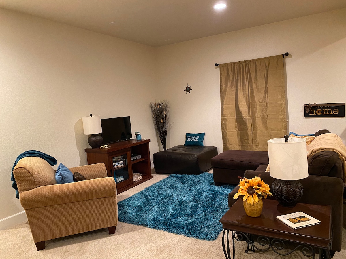 A cozy living area features a dark sofa adjacent to a soft ottoman, complemented by a light brown armchair. A plush blue rug anchors the space, while a simple coffee table holds decorative items and a lamp. Warm light illuminates the muted walls, enhancing the welcoming feel.