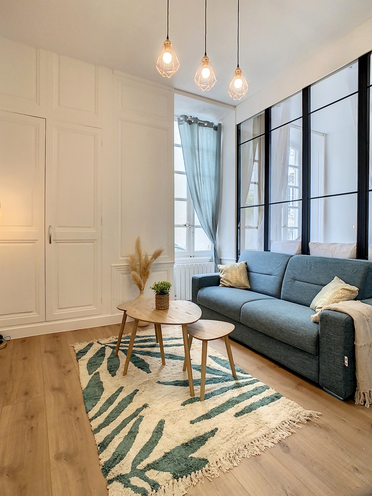 A cozy living area is presented, featuring a blue sofa complemented by decorative cushions. Natural light filters through large windows adorned with soft curtains. A pair of small wooden tables sits on a patterned rug, while a potted plant adds a touch of greenery to the space.