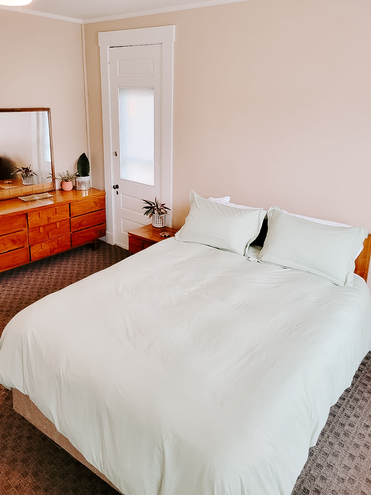 A cozy bedroom is highlighted by a large bed dressed in a soft green duvet. A dresser with wooden finishes stands against the wall, accompanied by decorative plants. Natural light enters through a nearby window, enhancing the inviting atmosphere.