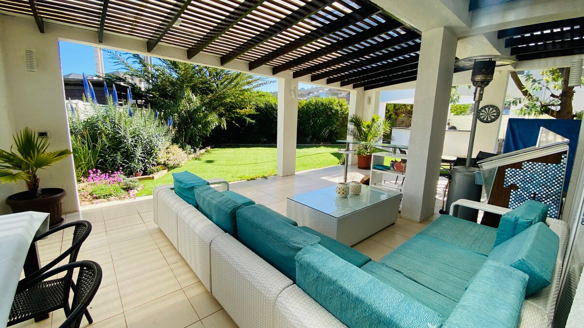 A covered outdoor area features a spacious sectional sofa with turquoise cushions, positioned beside a glass table. Lush greenery and colorful flowers surround the open space, providing a serene view. The patio is illuminated by natural light filtering through the overhead structure.