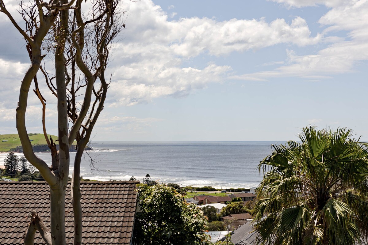 An expansive coastal view is presented, showcasing tranquil blue waters and gentle waves. Lush greenery and palms are framed in the foreground, complementing the scenic landscape. The sky features soft clouds, allowing light to enhance the serene atmosphere of the surroundings.