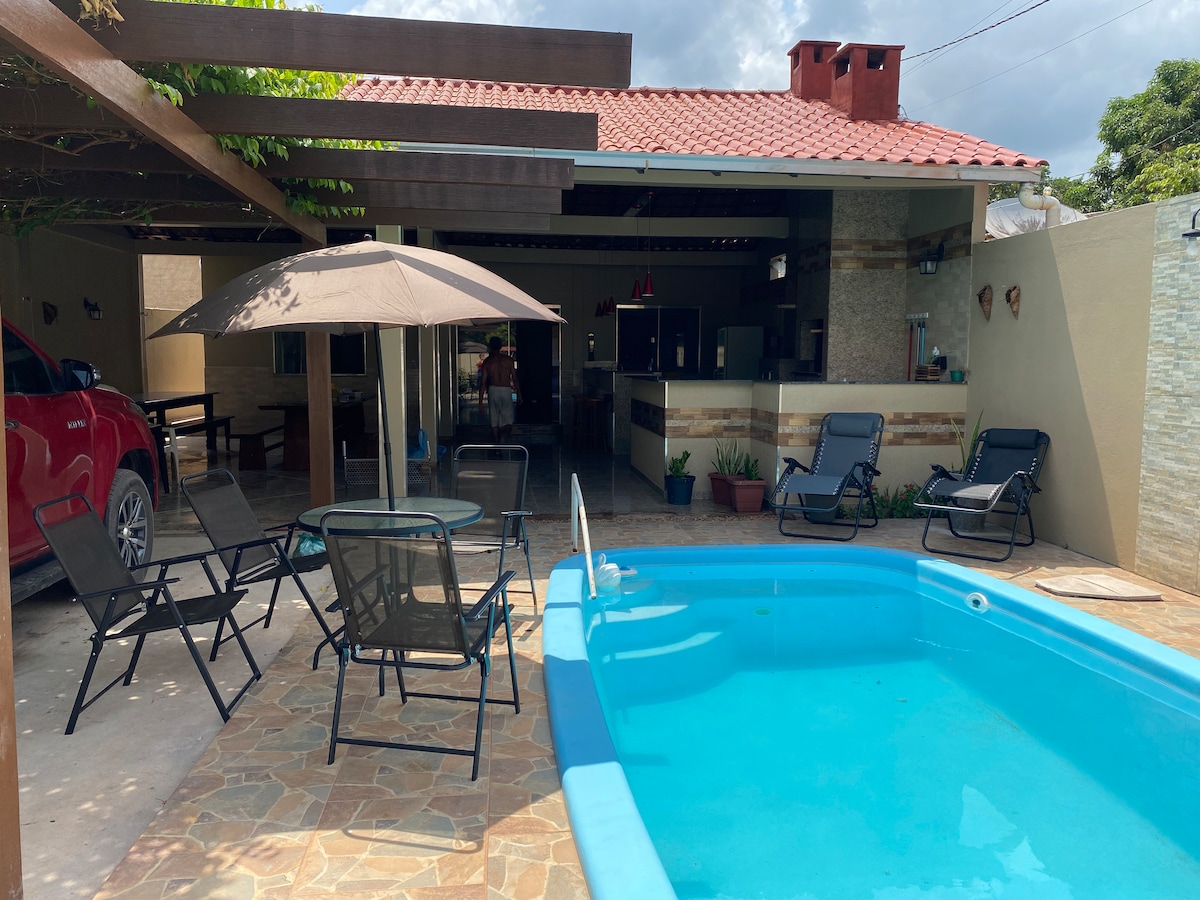 An outdoor area features a refreshing pool surrounded by a stone patio. Several lounge chairs are positioned nearby, and a shaded table with an umbrella offers a spot for relaxation. The backdrop includes a sturdy structure with a tiled roof and ample natural light.
