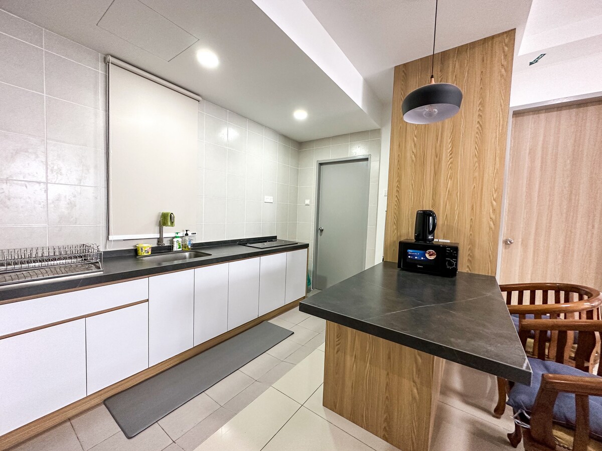 A modern kitchen space features a combination of sleek cabinetry and a dark countertop. Essential appliances, including a kettle and a microwave, are visible. Natural light illuminates the area, and a comfortable seating arrangement is provided nearby.