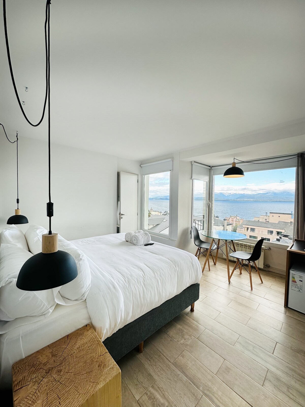 A bright guest room features a spacious bed with crisp white linens. Large windows offer a panoramic view of the surrounding landscape. Two wooden chairs and a small table are positioned near the windows, while a stylish nightstand with hanging lights adds to the minimalist decor.