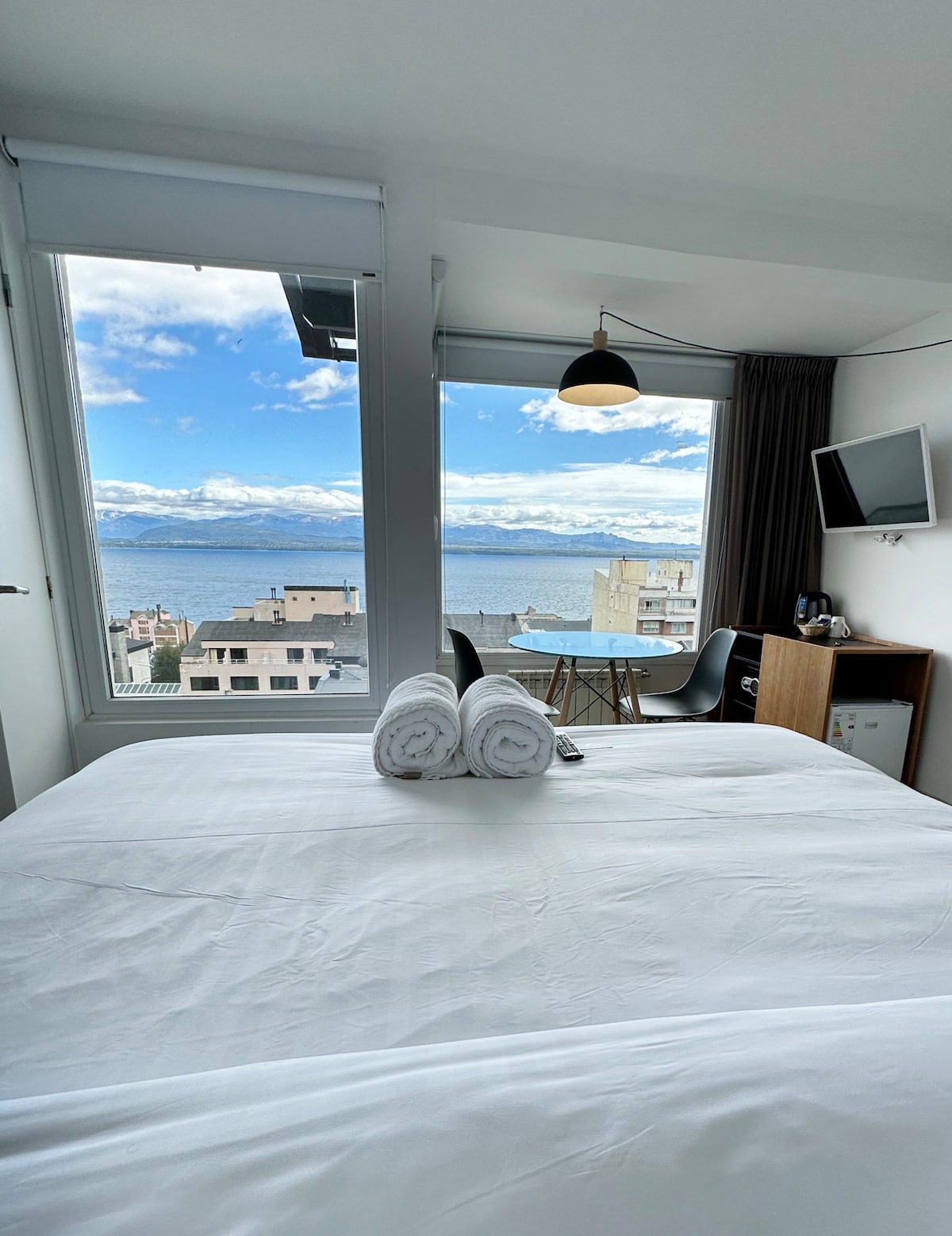 A spacious room features a bed with crisp white linens and neatly rolled towels placed on top. Large windows offer a panoramic view of the lake and surrounding mountains, allowing natural light to fill the space. A television and a small desk can also be seen.