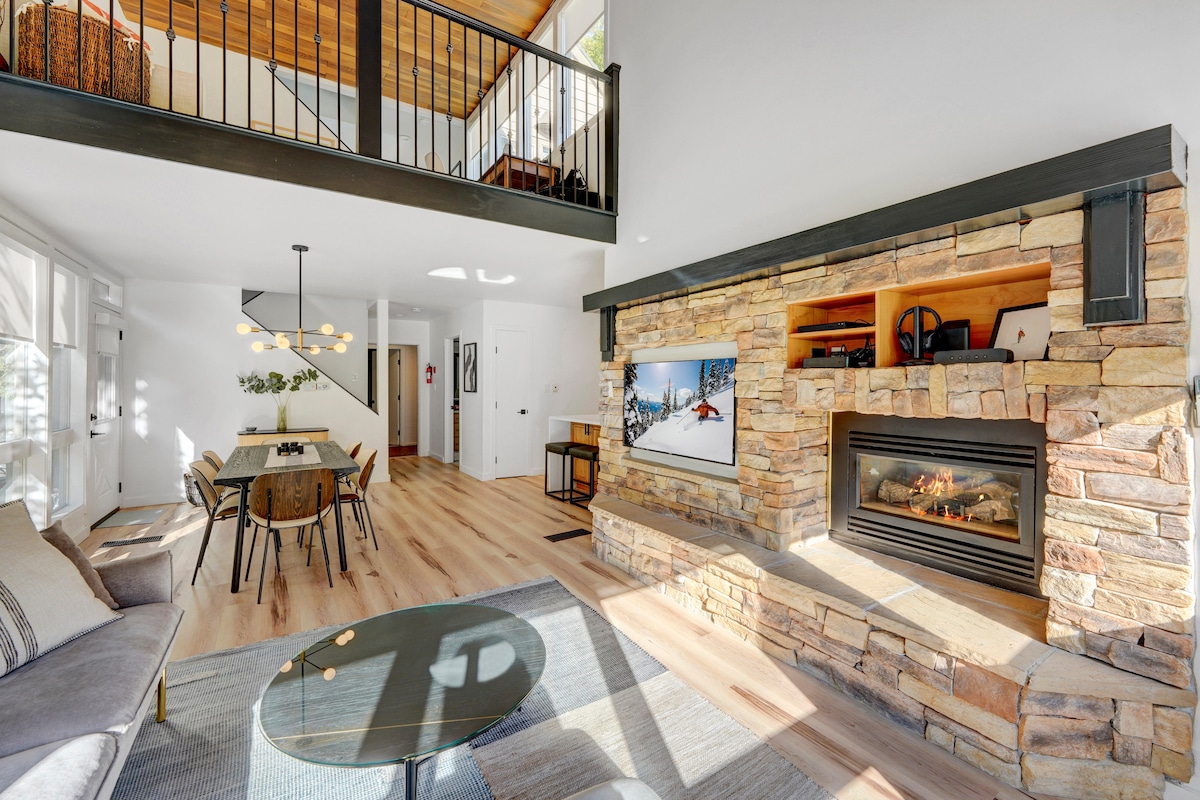 An open living area features a stone fireplace, surrounded by a TV and shelves. Large windows allow natural light to flood the space. The dining area showcases a round table with stylish chairs, while a loft overlooks the setting. Modern furnishings create a welcoming atmosphere.