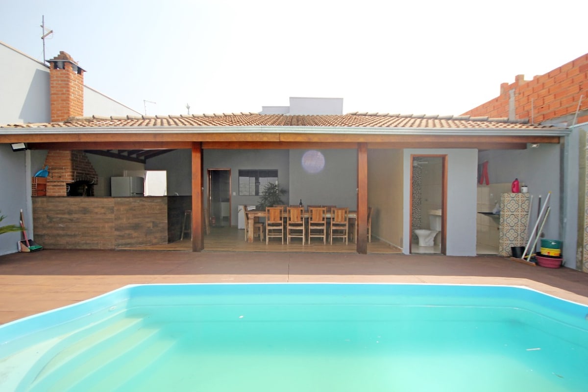 A serene outdoor area is highlighted by a clear blue pool, accompanied by a spacious wooden deck. A covered dining space is visible, featuring a large table surrounded by multiple chairs. The background includes a kitchen area with modern finishes and a neutral color palette.