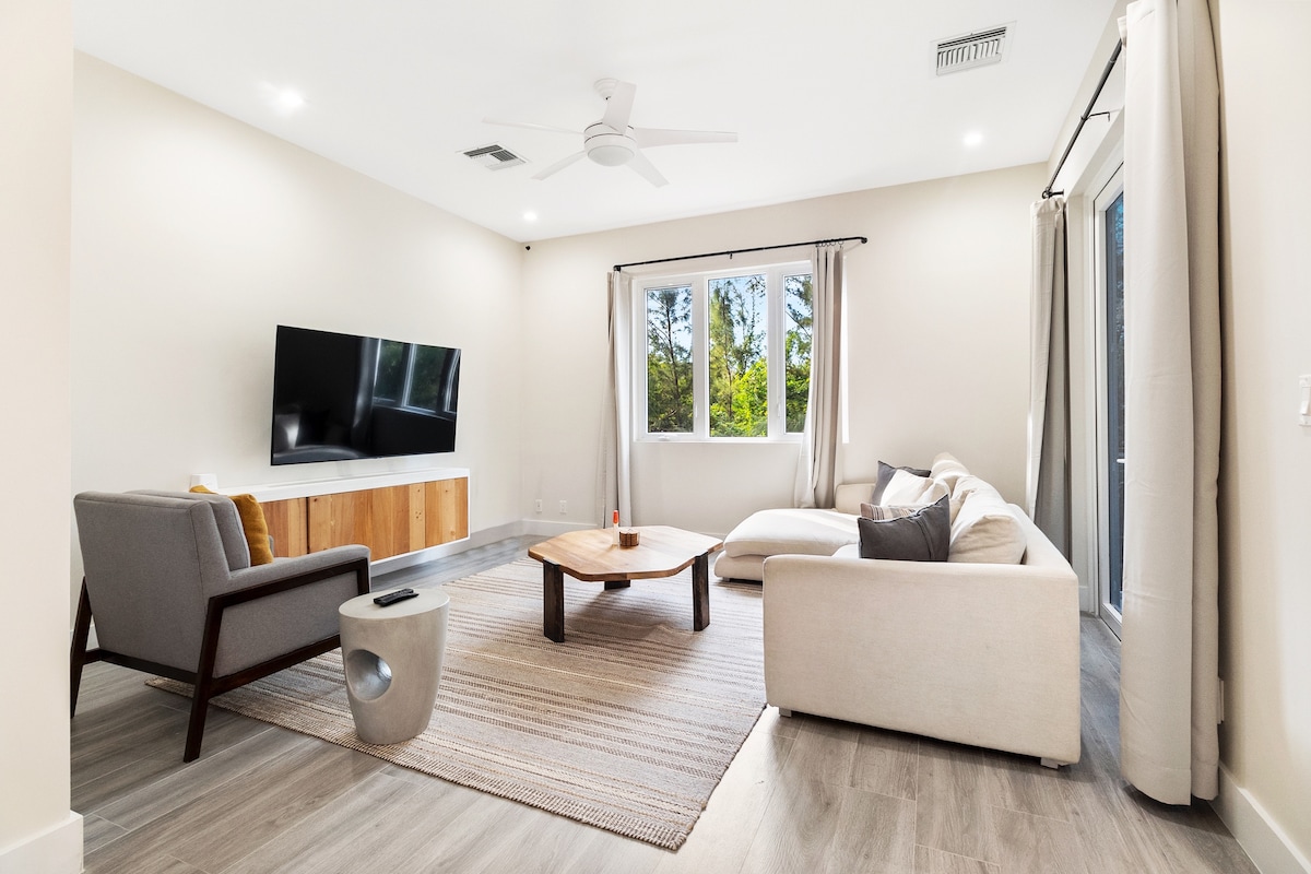 A modern living area features a large flat-screen television mounted on the wall, complemented by a wooden media console. A plush sectional sofa and an accent chair surround a natural wood coffee table, placed on a soft area rug. Large windows allow natural light to illuminate the space.
