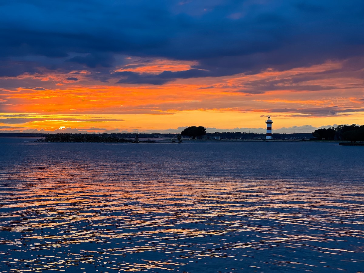 A serene lakeside view features a colorful sunset reflecting on the calm waters. A lighthouse stands in the distance, framed by a blend of blue and orange skies. The horizon is marked by silhouettes of trees and a rocky shoreline, creating a peaceful natural scene.