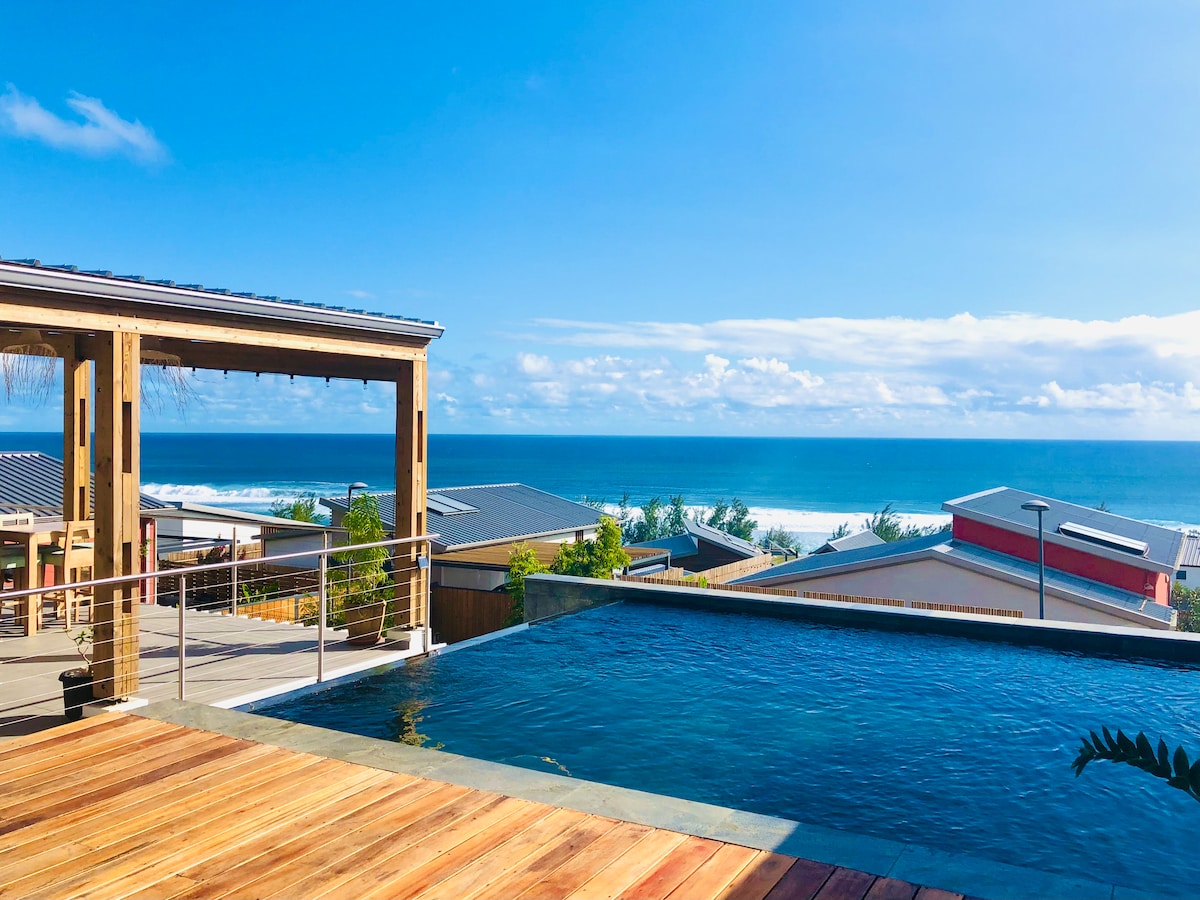 An outdoor area features a spacious infinity pool that blends into the ocean view. The deck, made of warm wood, provides space for relaxation. A dining area with a shaded pergola is visible, set against the backdrop of a clear blue sky and distant coastal scenery.