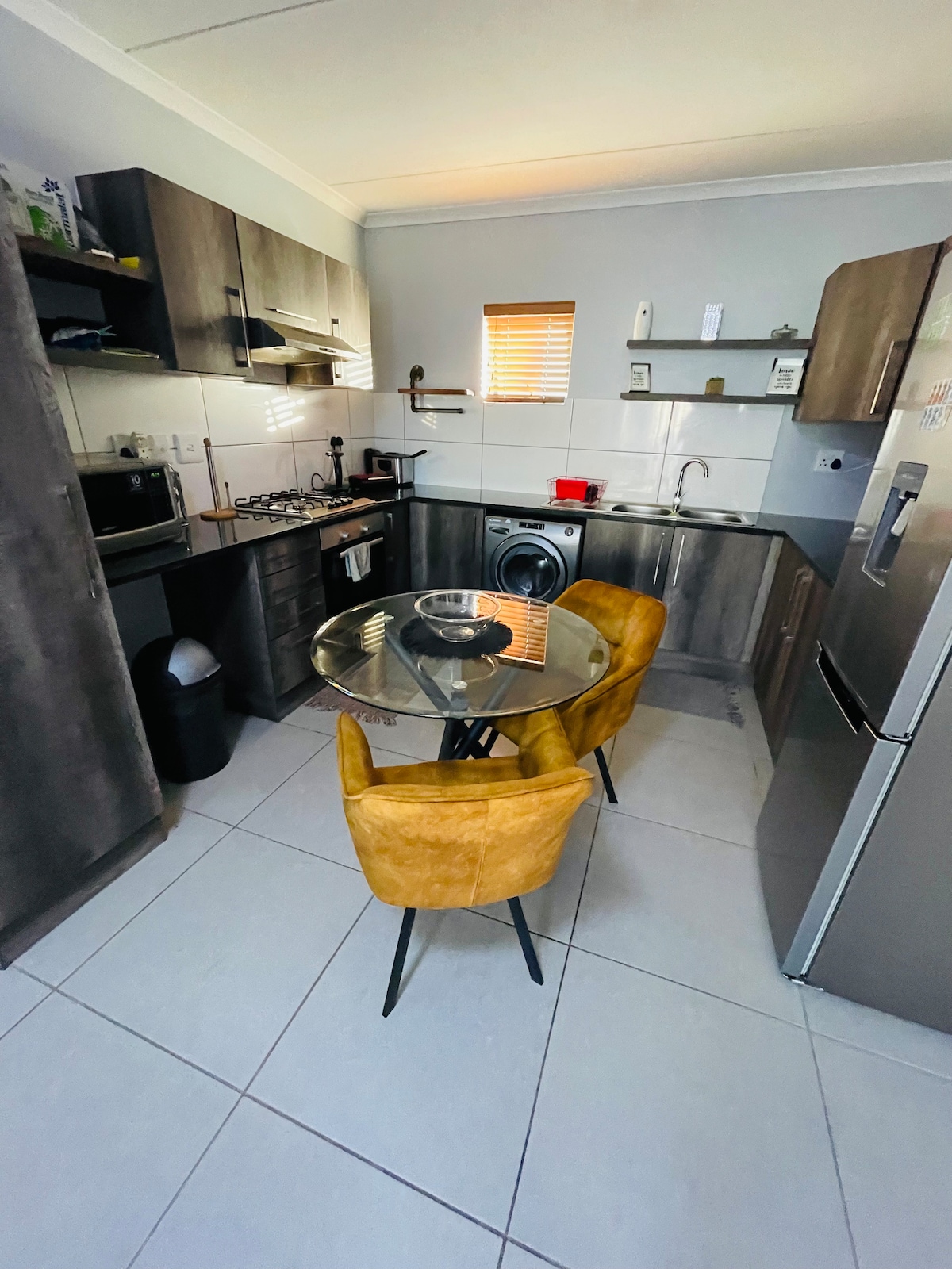 The kitchen features a modern arrangement with dark cabinetry and a glass-top dining table surrounded by yellow chairs. Stainless steel appliances, including a refrigerator and a gas stove, are visible, along with a small washing machine. Natural light filters through a window, enhancing the space.