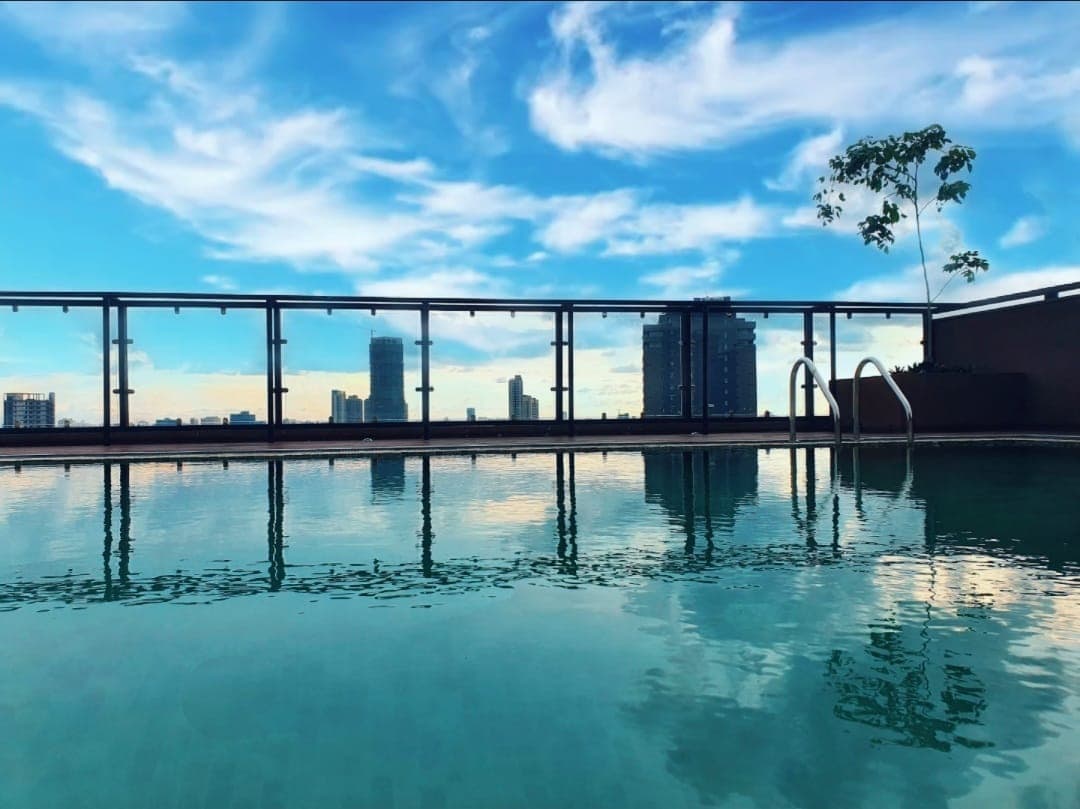 A rooftop swimming pool reflects the blue sky and clouds, complemented by views of the surrounding cityscape. The pool features a smooth edge, with a ladder leading into the water, and a small tree is visible along the perimeter.