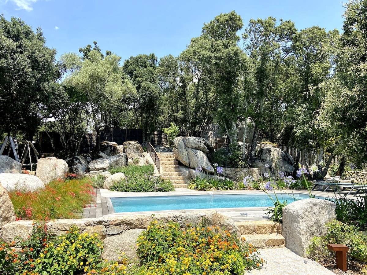 A private heated pool is framed by natural rock formations and vibrant greenery. Lush plants and flowers surround the pool area, while stone steps lead to an elevated spot. Shade is provided by various trees, creating a serene outdoor space for relaxation.