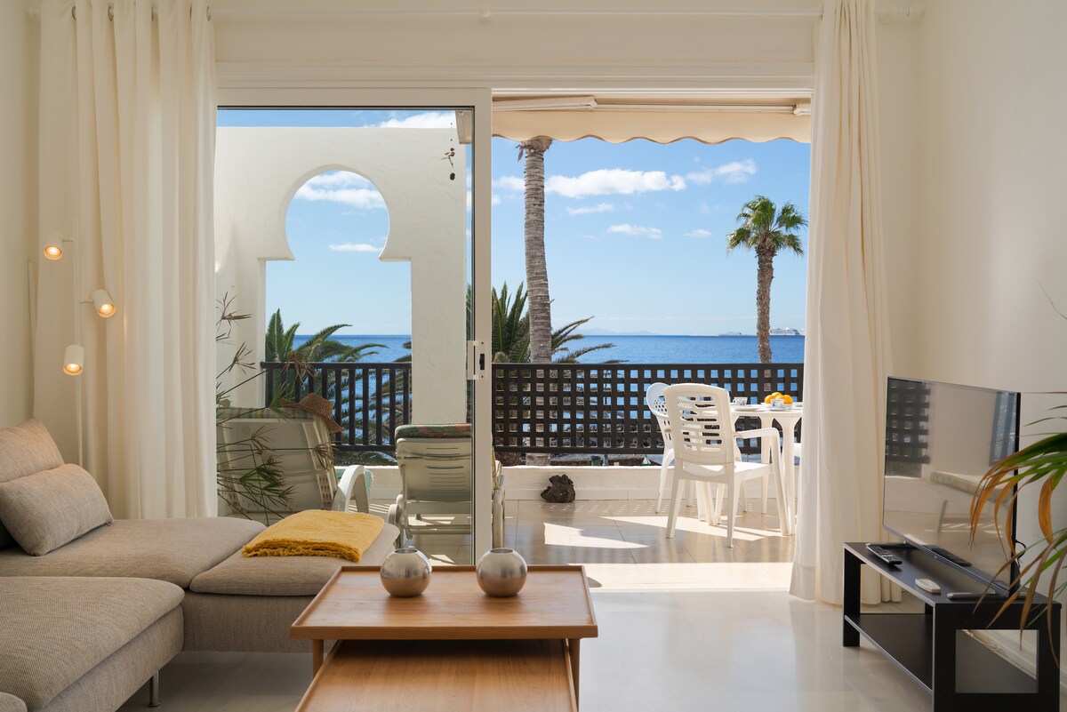 Natural light floods a living area featuring a gray sectional sofa and wooden coffee table. A view of palm trees and the ocean is visible through large sliding glass doors, which open onto a terrace with outdoor seating and a small dining set.