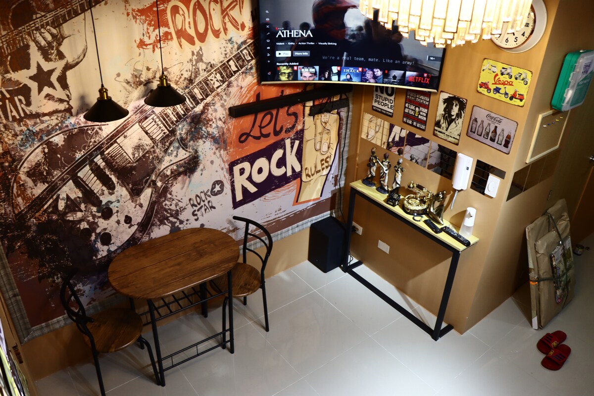 A dining area is viewed from above, featuring a wooden table surrounded by two black chairs. The walls are adorned with colorful rock music-themed decor, showcasing band posters and memorabilia. A television is mounted above a sleek console, enhancing the lively ambiance.