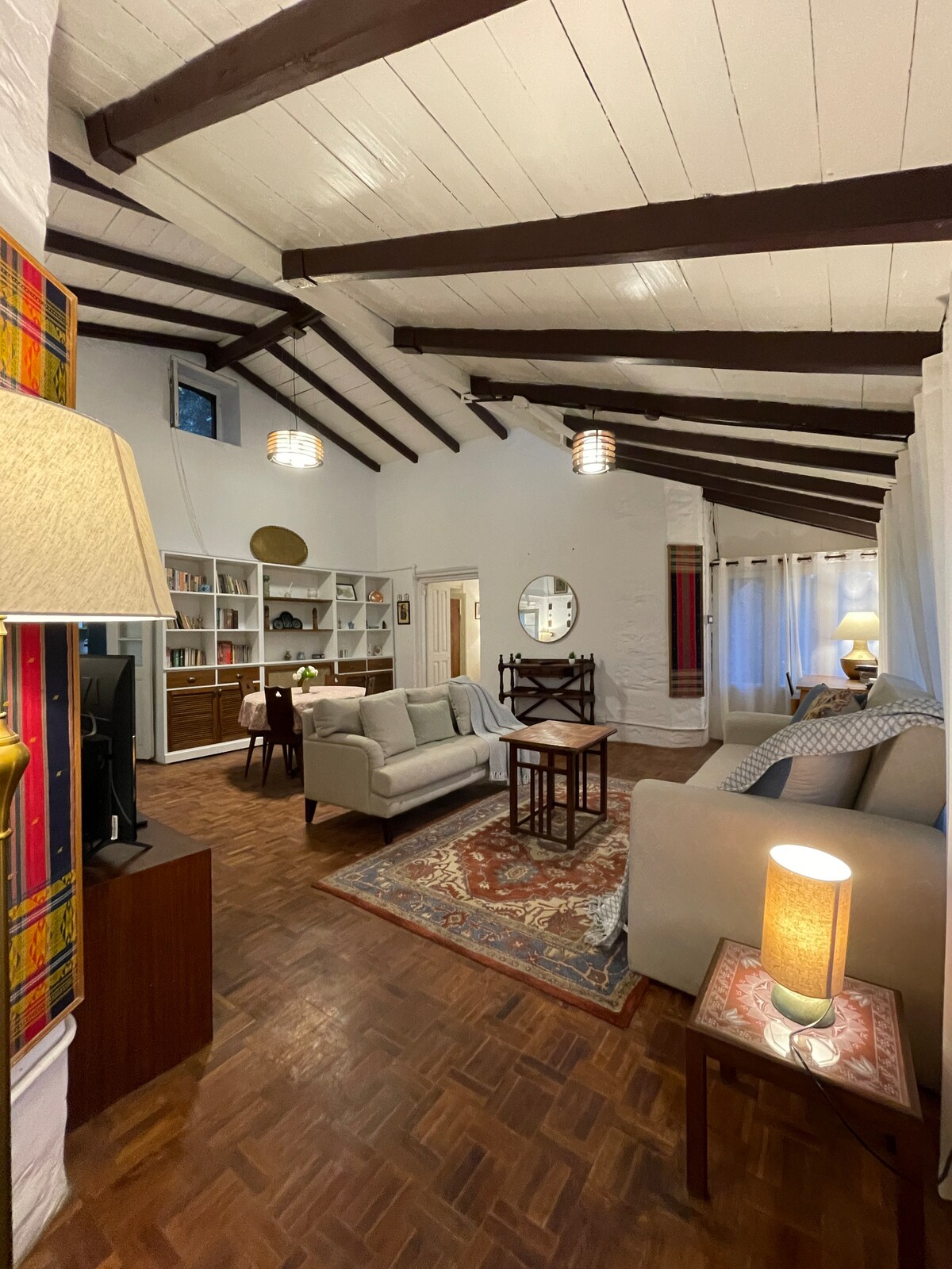A spacious living area is highlighted by high wooden beams and soft, natural light. Comfortable sofas are arranged alongside a decorative rug, with a small dining space visible. A bookshelf and mirrors add depth, while warm accents create a cozy ambiance.