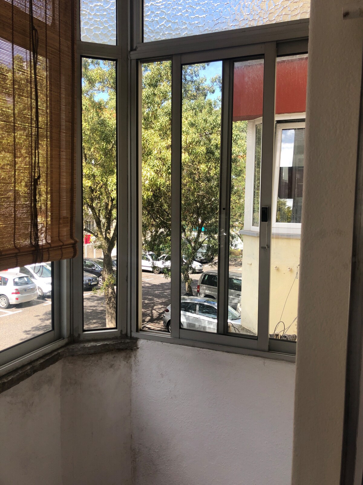 A corner window with multiple panels allows natural light to fill the room. Outside, trees provide greenery, and parked cars can be seen along the street. A bamboo shade partially covers the window, adding a touch of warmth to the space.
