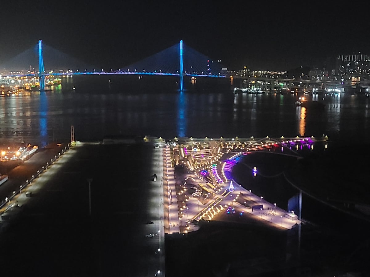 The vibrant night scene captures a panoramic view of the illuminated Busan Harbor Bridge, featuring bright blue lights against the dark sky. The waterfront walkways are adorned with colorful lights, reflecting off the tranquil waters, highlighting the lively atmosphere of the surrounding city.