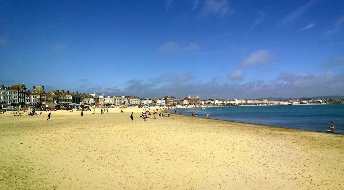 A wide sandy beach stretches along the shoreline under a clear blue sky. The gentle waves lap at the shore, while people enjoy various activities, including walking and relaxing on the sand. Colorful beach huts and buildings line the background, adding to the vibrant coastal scene.