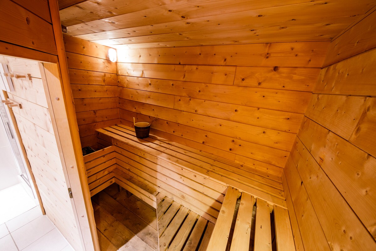 A wooden sauna is presented, featuring warm-toned wood paneling and benches arranged along two sides. A small bucket is positioned on one of the benches, and soft lighting creates a calming ambiance within the enclosed space.