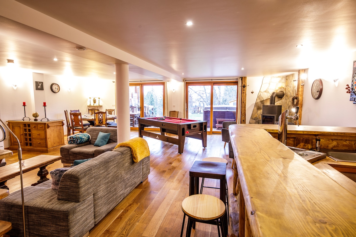 A spacious living area features comfortable seating arrangements and wooden furnishings. Large windows provide ample natural light and views of the outdoor scenery. A pool table is positioned centrally, while a wooden bar creates an inviting space for gatherings.